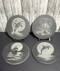 Four round coasters with black and white nautical designs displayed on a dark surface against a rustic wood plank backdrop. The coasters feature illustrations of dolphins, killer whale, palm trees, a ship, and a full moon over water.