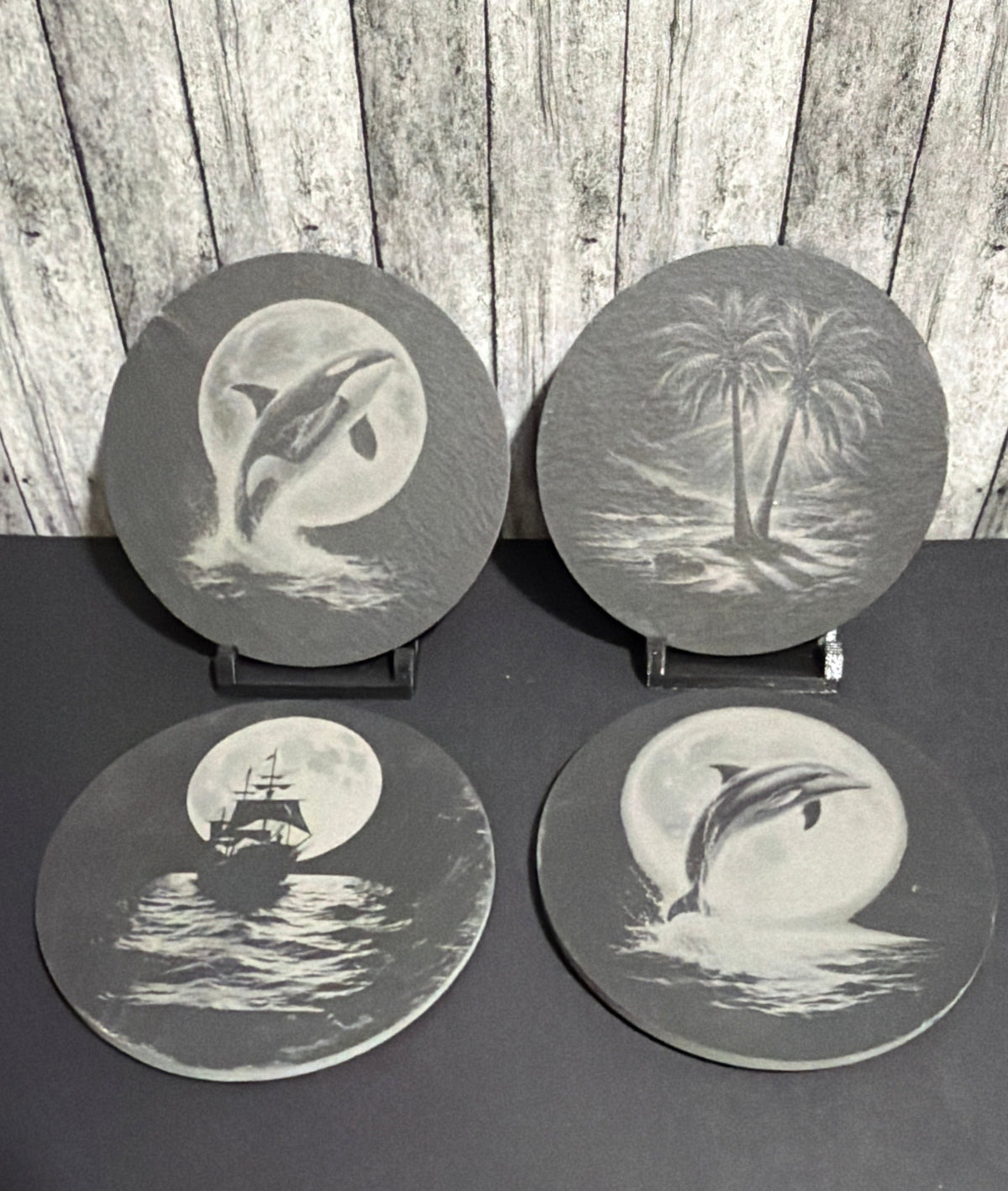 Four round coasters with black and white nautical designs displayed on a dark surface against a rustic wood plank backdrop. The coasters feature illustrations of dolphins, killer whale, palm trees, a ship, and a full moon over water.